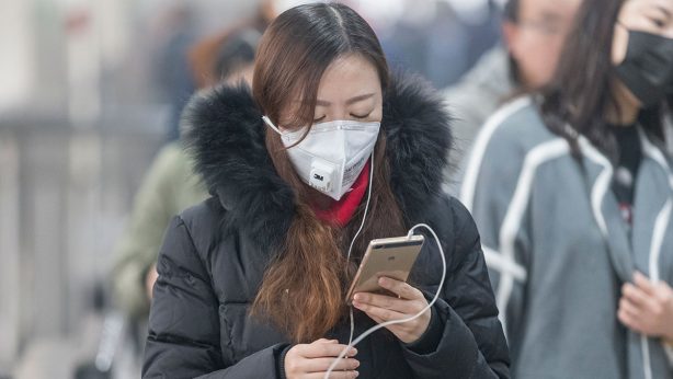 Woman wearing a face mask and winter coat using a smartphone outdoors.